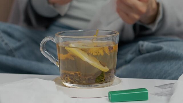 A woman sits in bed, checking her temperature while sipping herbal tea. She is dealing with a cold and tries to recover from her symptoms and fever.