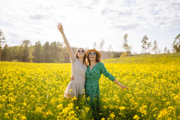 Fototapeta premium Happy girlfriends in dresses are having fun in a yellow field in the sun's rays. Two young women are strolling, spending time together outdoors. Concepts of friendship and fashion.