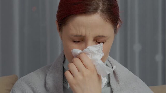 A woman sits on the couch feeling unwell. She is wrapped in a blanket and uses tissues to help with her symptoms. She looks tired and is trying to rest.