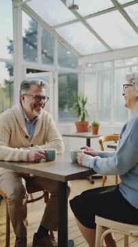 Elderly couple enjoying a peaceful conversation over coffee in a bright, plant-filled caf&eacute;, vertical shot