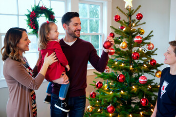 Naklejka premium Caucasian man holding Christmas ornament while smiling at Caucasian young adult woman holding child, teenage boy standing near decorated Christmas tree during holiday celebration