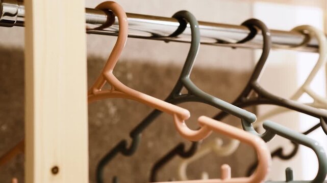 A close-up of a row of plastic coat hangers in pastel shades (beige, grey, white, peach) hanging neatly on a metal rod. Soft lighting creates a warm, minimalist atmosphere.
