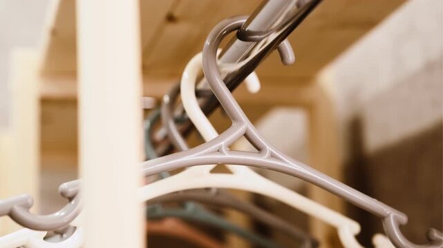 A close-up of a row of plastic coat hangers in pastel shades (beige, grey, white, peach) hanging neatly on a metal rod. Soft lighting creates a warm, minimalist atmosphere.