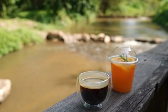 Black coffee and orange juice in a glass on wooden sheet with blurred by the river background.