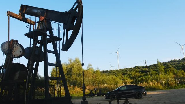 Oil pumpjack stands beside car with wind turbine above shrubs. Counterweight rotates while walking beam dips and lifts with steady rhythm