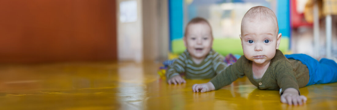 Two baby twin brothers are crawling on a bright yellow floor. One twin is closer to the camera while the other is in the background. The room has a playful environment. Empty space for text