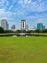 Obraz premium Jakarta cityscape viewed from the Monas area, featuring wide green lawns in the foreground and modern high rise buildings under a bright blue sky with scattered clouds.