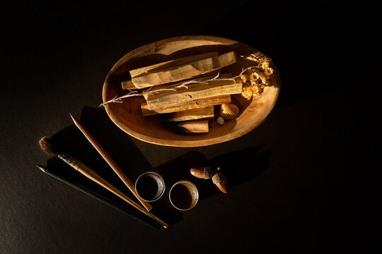 Table vue du dessus, composition de mat&eacute;riel de dessin, pinceau, crayon, godets d'aquarelle brun et marron, coupelle bois avec Palo Santo, fleur s&eacute;ch&eacute;e, gland brun, fond brun fonc&eacute;, lumi&egrave;re contrast&eacute;e