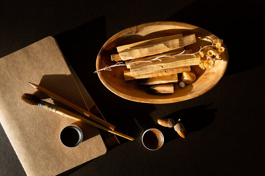 Vue du dessus d'une table avec du mat&eacute;riel de dessin, carnet, pinceau, crayon, godets d'aquarelle brun et marron, plateau bois avec Palo Santo, fleur s&eacute;ch&eacute;e, gland brun, fond fonc&eacute; et rayon de lumi&egrave;re