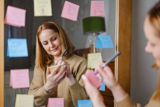 Woman writing positive affirmations on sticky notes in front of mirror