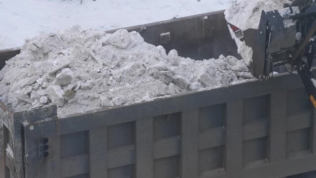 Specialized equipment performs snow removal in the city after a heavy snowfall. A front loader dumps a full bucket of dirty ice and snow into the body of a dump truck