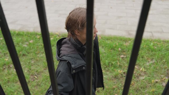 pensive person behind metal fence with wet hair and headphones, standing on urban sidewalk under overcast sky, contemplative expression, muted coat with rain droplets, solitary mood and subtle tension