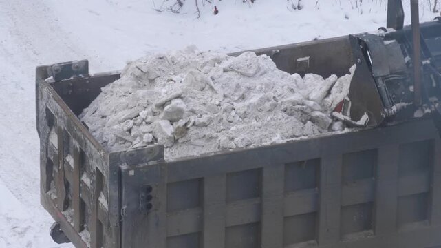 A front loader fills the bed of a dump truck with dirty compacted snow during winter road clearance. Heavy machinery working to remove snow from city streets after a blizzard