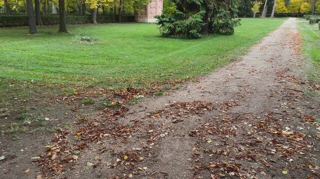 Images in the Royal Gardens of Aranjuez, a dirt path and freshly cut grass, the camera rises, revealing a brick obelisk, partially surrounded by trees, part of the gardens' historic hydraulic system.