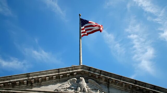 Flag flies above courthouse pediment. Flagpole rises from classical building roof. Stone eagle adorns pediment facade. Government architecture is emphasized against sky. Patriotism felt in frame view.