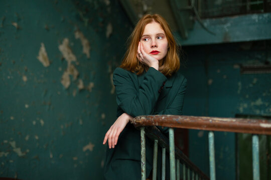 Red-haired teenager in green jacket leaning on old staircase railing indoors