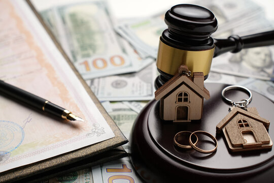 Property Division in Divorce and Marital Law Concept. Gold wedding rings, wooden house model, and a judge gavel on legal paperwork and US currency. Division, alimony, and real estate legal disputes