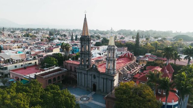 Aerial view of the Immaculate Conception Church, its spires piercing the sky amidst a tapestry of red-tiled roofs and verdant trees, Chapala, Jalisco, Mexico.