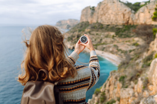 View from behind. Hiker in hat stands on edge of cliff, holding compass, overlooking sea. Young woman with compass on hiking trail enjoys scenery and feels sense of freedom. Navigation concept.
