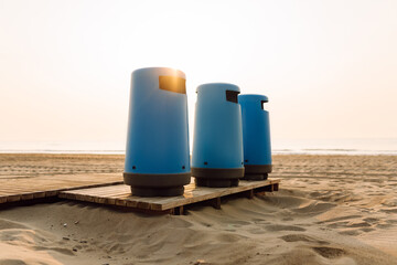 Naklejka premium Three blue trash bins stand on a wooden platform on the beach sand. Trash containers on a public beach in the sunset. The concept is ecology, cleanliness, and nature.