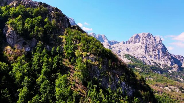 Aerial drone view of Theth National Park, Albania, alpine mountains and rocky hills with pine trees, village and hiking switchback trail in summer, pristine nature, clear daylight, July 2023
