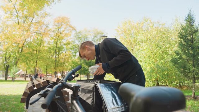 Delivery rider loading groceries onto bike, leaning over rear rack, securing panniers and boxes on sunny autumn day, trees and pathway in background, casual jacket, hands fastening straps, bicycle