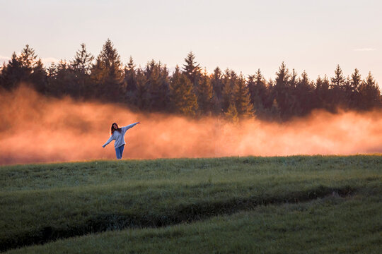 Woman with arms outstretched standing in front of haze on a meadow at sunset