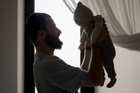 Father holds baby near window in warm, natural light capturing tender family moment