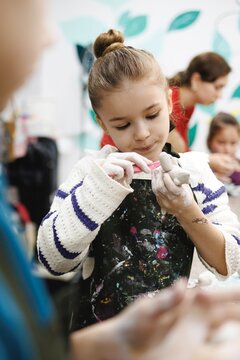 Young girl focused on shaping clay at a creative workshop with bright colors and other children around her