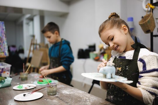 Young girl smiles as she paints a small sculpture in a bright art studio with a boy working nearby