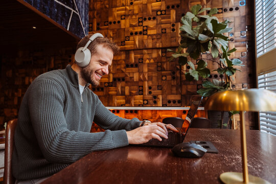 Man smiling wearing headphones while typing on a laptop computer working remotely from a modern coffee shop.