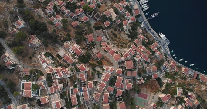 Aerial view of the clustered red-roofed buildings cascading down the hillside towards the deep blue sea filled with boats, Simi, Rhodes, Greece.