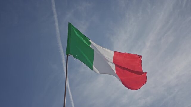 Italian flag waves on a pole above a street with green white and red fabric fluttering in blue sky; pride.