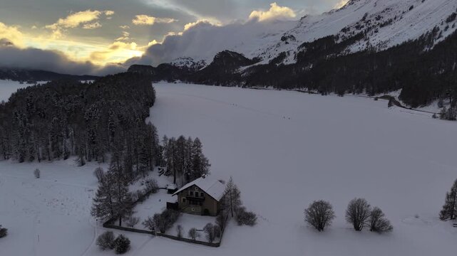 Aerial view of snow-covered landscape with trees and houses under a cloudy sky, creating a serene winter scene, Sils im Engadin/Segl, Switzerland.