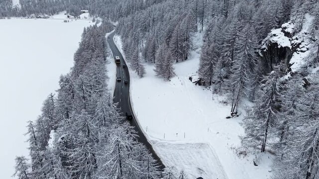 Aerial view of cars traveling along a winding road cutting through a snow-covered forest next to a frozen lake, Sils im Engadin/Segl, Grigioni, Switzerland.