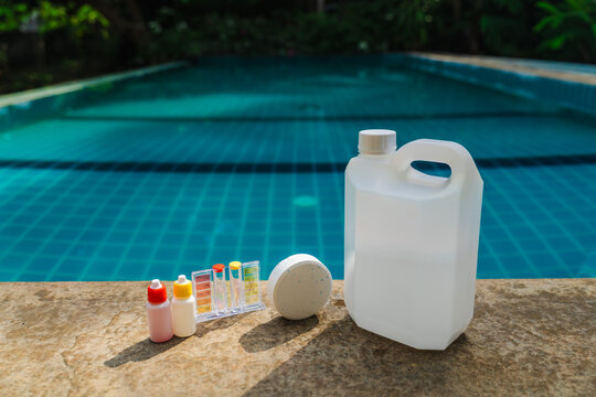 Pool care essentials including chlorine tablet, water testing kit and liquid chemical bottles arranged on stone ledge beside outdoor swimming pool, tropical poolside maintenance concept
