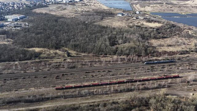Aerial view of trains moving along tracks near factories and bare trees, creating a stark contrast between industrial and natural landscapes, Wittelsheim, Grand Est, France.
