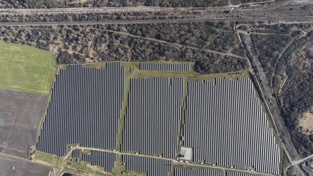 Aerial view of a vast solar farm, its dark panels contrasting with the adjacent green field and bare trees, Wittelsheim, Grand Est, France.