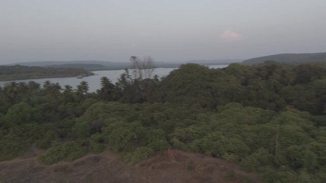 360-degree aerial view of Querim Beach, North Goa, showcasing a long sandy coastline with rocky outcrops, rows of trees, and the Tiracol River inlet under soft winter evening light