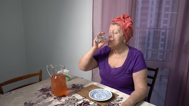 Elderly alone woman drinks compote from a glass at home. Pour a drink from a carafe. Grandma