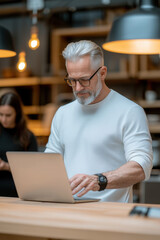 Mature man gray hair beard glasses white sweater smartwatch laptop coworking space modern office warm light with focus professional mood in workspace