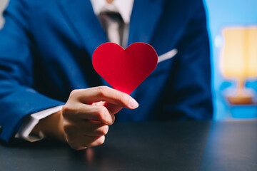 Love concept. Red heart shape held by person in blue suit symbolizing love and affection with blurred background and warm lighting