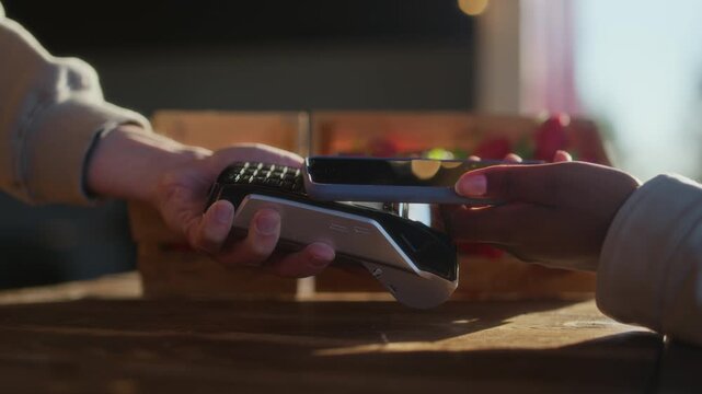 Closeup of unrecognizable buyer handing smartphone to farm vendor during fresh strawberry purchase at rustic outdoor market stall