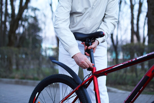A person in a white jacket adjusts the bike saddle height on a red bicycle, focused, practical outdoor maintenance amid bare trees and soft daylight.