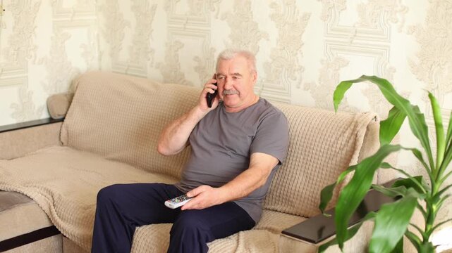 An older man with a mustache sits on a beige sofa, talking on a mobile phone, calm expression, patterned wallpaper, plant nearby.