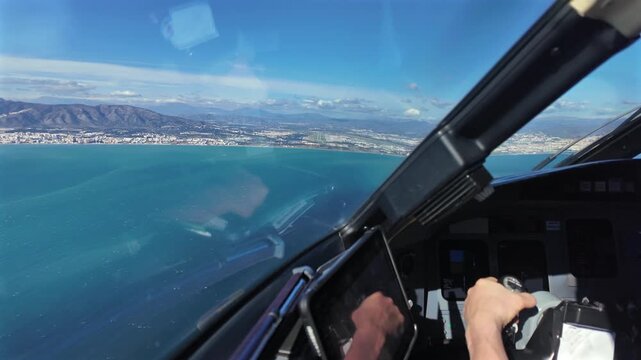 Hand of a caucasian pilot taking the manual control of a jet airplane grasping the yoke, while overflying the sea with Malaga airport (AGP) runway ahead, in a sunny and windy day.