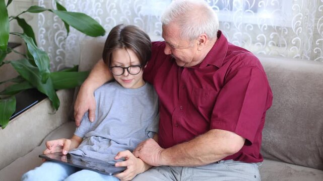 An older man embraces a boy on the sofa as they share a tablet, smiling, connected, in a tender intergenerational moment bathed in soft daylight.