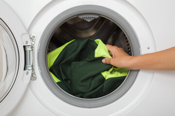 Young adult woman hand holding and unloading dark green workwear with reflective elements from opened white washing machine. Clothes care. Closeup. Front view.