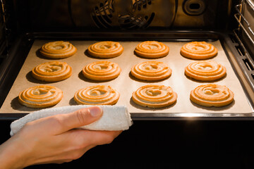 Young adult woman hand with towel holding tray with fresh dry shortbread cookies in oven. Closeup. Baking sweet snack at home. Front view.