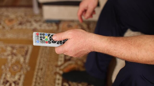 Close up of hands gripping a TV remote on a couch, thumb actively pressing buttons, casual home setting.
